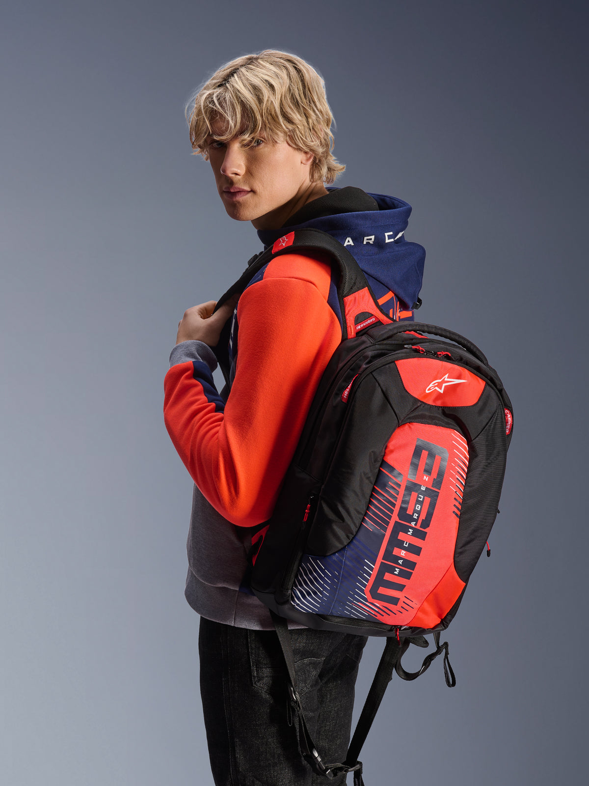 Mochila Alpinestars MM93 City Hunter V2, negra con detalles en rojo flúor y azul marino, con gráficos de la firma MM93 de Marc Márquez, combinada con una sudadera deportiva a juego, perfil aerodinámico estilizado para trayectos en moto
