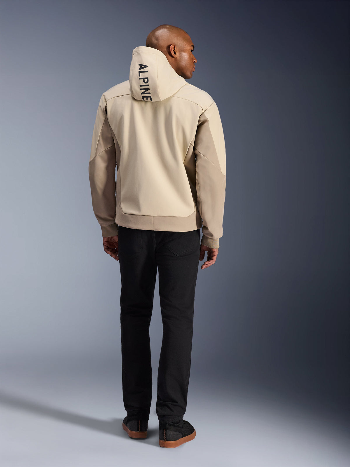 Sudadera con capucha Alpinestars Radium Tech, Mastic Aluminum, vista trasera que muestra el texto vertical 'ALPINE' en la capucha, chaqueta técnica de moto urbana con capa exterior 3L cortavientos y paneles color crema, combinada con pantalones negros