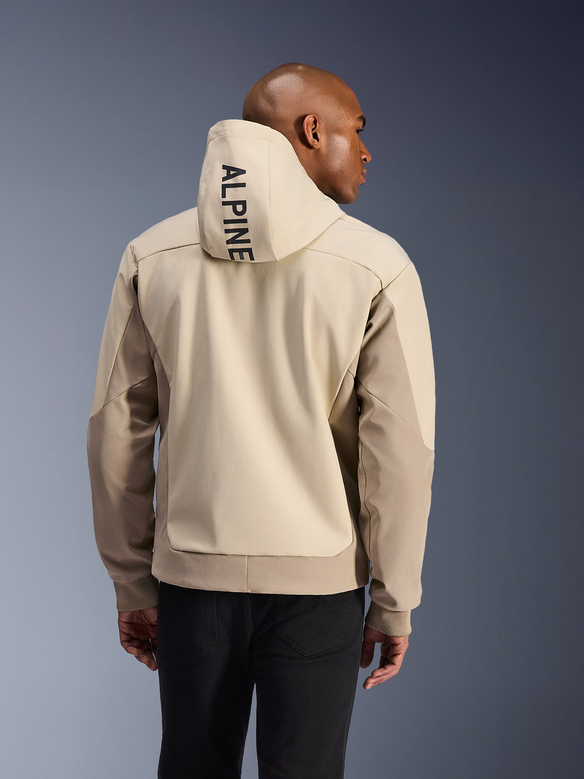 Sudadera con Capucha Alpinestars Radium Tech, vista trasera de un hombre usando la sudadera técnica Mastic Aluminum, con el texto ALPINE en vertical en la capucha, paneles en beige claro y arena, puños acanalados, chaqueta de conducción urbana para motocicleta