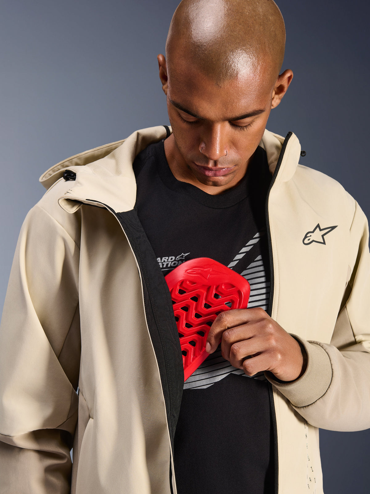 Sudadera con capucha Alpinestars Radium Tech en Mastic Aluminum, chaqueta de moto urbana, modelo mostrando el bolsillo interno para armadura con un protector de pecho Nucleon rojo, capa técnica cortaviento con diseño de sudadera casual y logotipo de Alpinestars.
