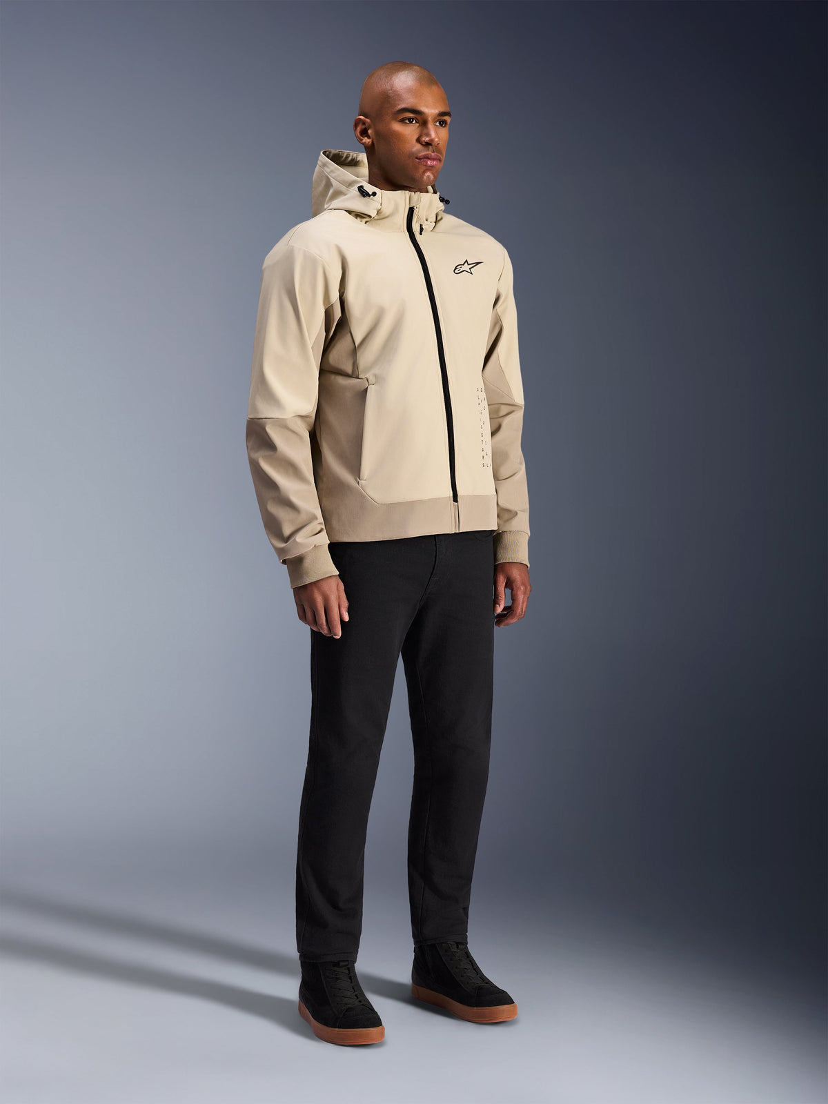Sudadera Alpinestars Radium Tech, chaqueta de moto urbana, color beige claro Mastic Aluminum, combinada con pantalones negros y zapatillas altas negras, con capa técnica cortavientos, cremallera frontal y capucha extraíble, diseño streetwear moderno para conducción urbana