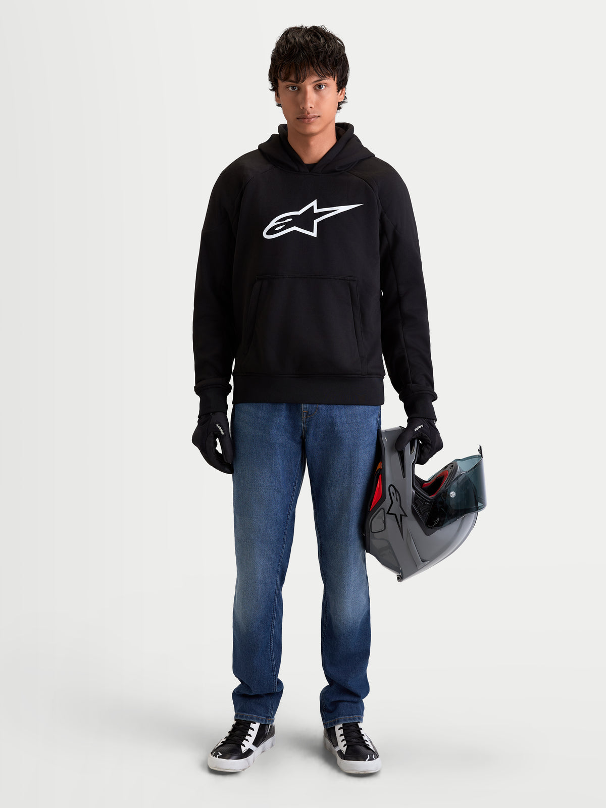 Alpinestars Chaqueta Con Capucha Ageless, sudadera de moto urbana para hombre en color negro con logotipo blanco en el pecho, vista frontal en modelo que sostiene un casco y guantes de moto