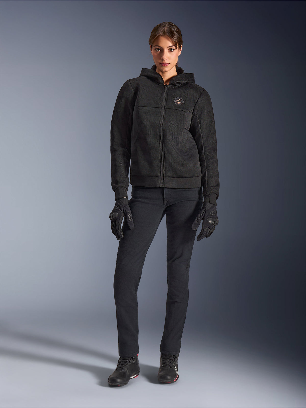 Modelo femenina luciendo la sudadera Alpinestars Stella Chrome Superairflow en color negro, combinada con pantalones técnicos de conducción y guantes negros, con construcción de malla 3D y logotipo en el pecho en tono cobre, ropa de motociclismo deportiva urbana para mujer.
