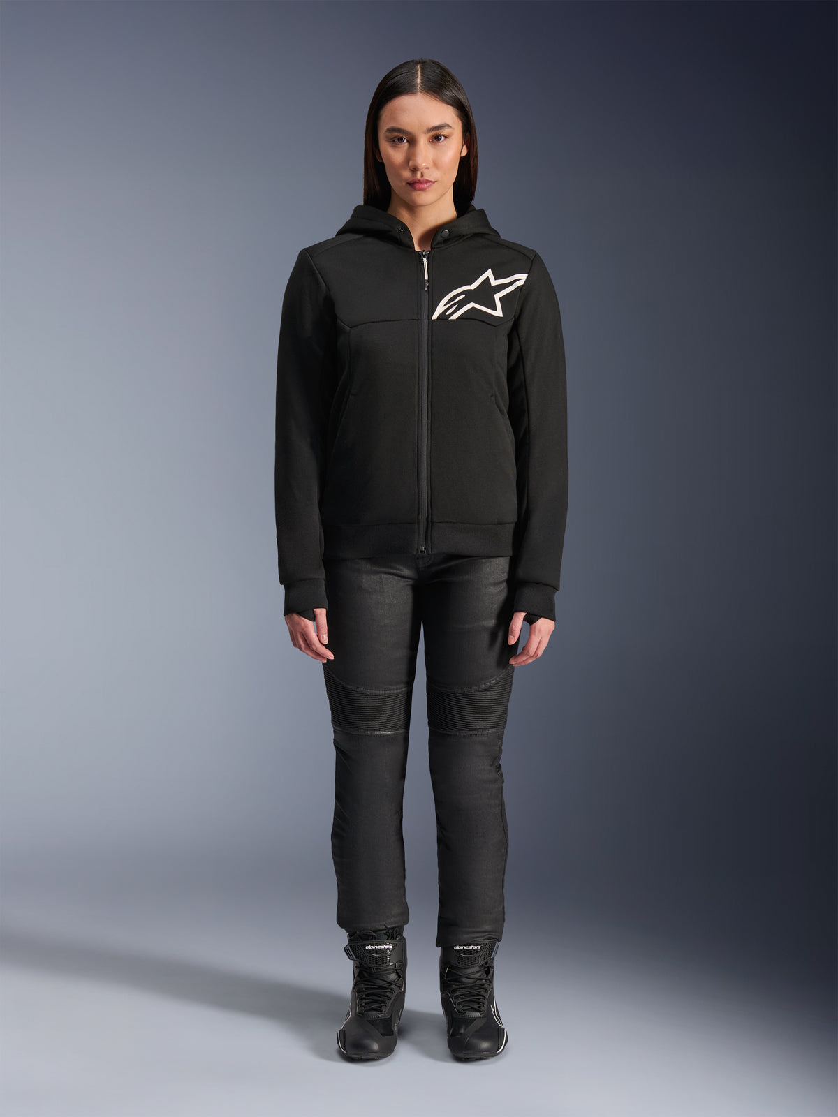 Mujer con la sudadera Alpinestars Stella Chrome V2 Sport, negra con el logotipo blanco de Astars en el pecho, combinada con pantalones técnicos de conducción y botas de color negro, diseño de ropa urbana para moto.