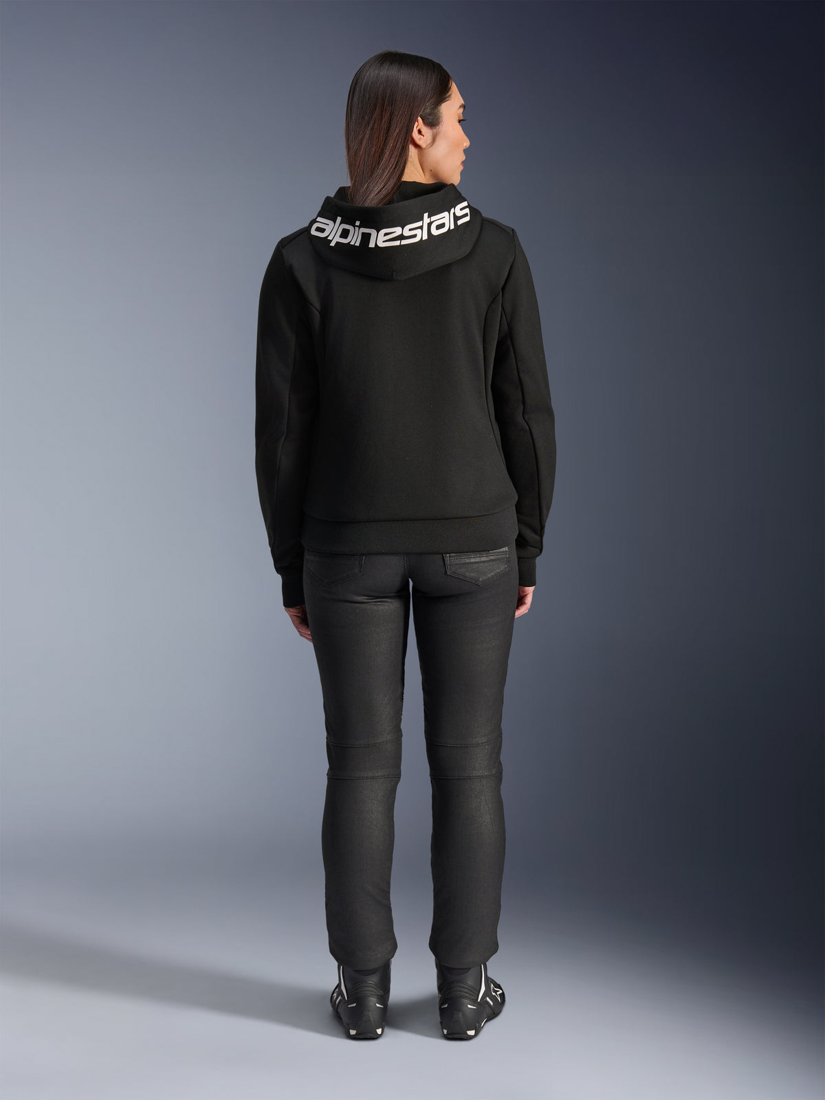 Vista posterior de una modelo con la sudadera Alpinestars Stella Chrome V2 Sport Hoodie en negro, con un logotipo blanco de Alpinestars destacado en la capucha, combinada con pantalones de conducción negros y botas de moto, equipamiento de moto deportivo urbano para mujer