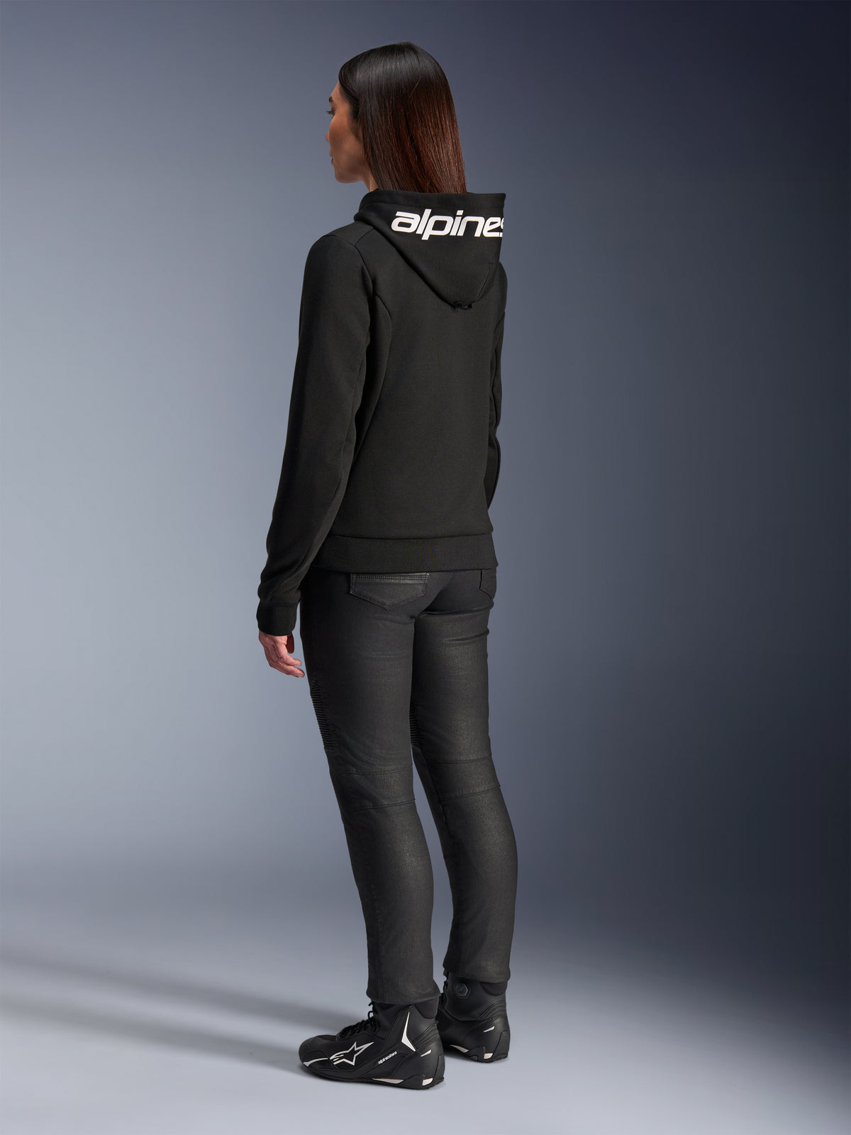 Mujer vistiendo la sudadera Stella Chrome V2 Sport Hoodie de Alpinestars, vista trasera, sudadera de moto negra con el logotipo blanco de Alpinestars en la capucha, combinada con pantalones técnicos de moto negros y calzado de moto negro, equipamiento urbano casual para desplazamientos.