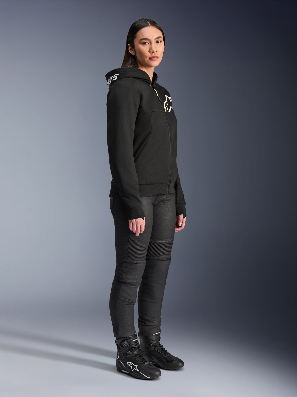 Mujer vistiendo la sudadera Alpinestars Stella Chrome V2 Sport Hoodie en negro, con logotipos de Alpinestars en blanco, combinada con pantalones de moto y botas negras, equipamiento de conducción urbana para mujer con un ajuste atlético casual
