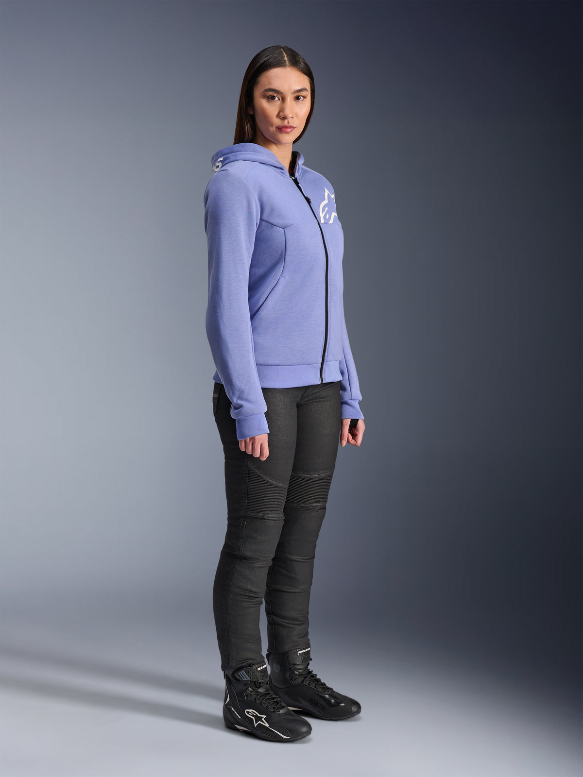 Alpinestars Woman Stella Chrome V2 Sport Hoodie, sudadera de moto en color azul violeta con el logotipo blanco de Alpinestars en el pecho, vestida por una modelo con pantalones de montar negros y botas de moto negras, ropa de conducción urbana para mujer