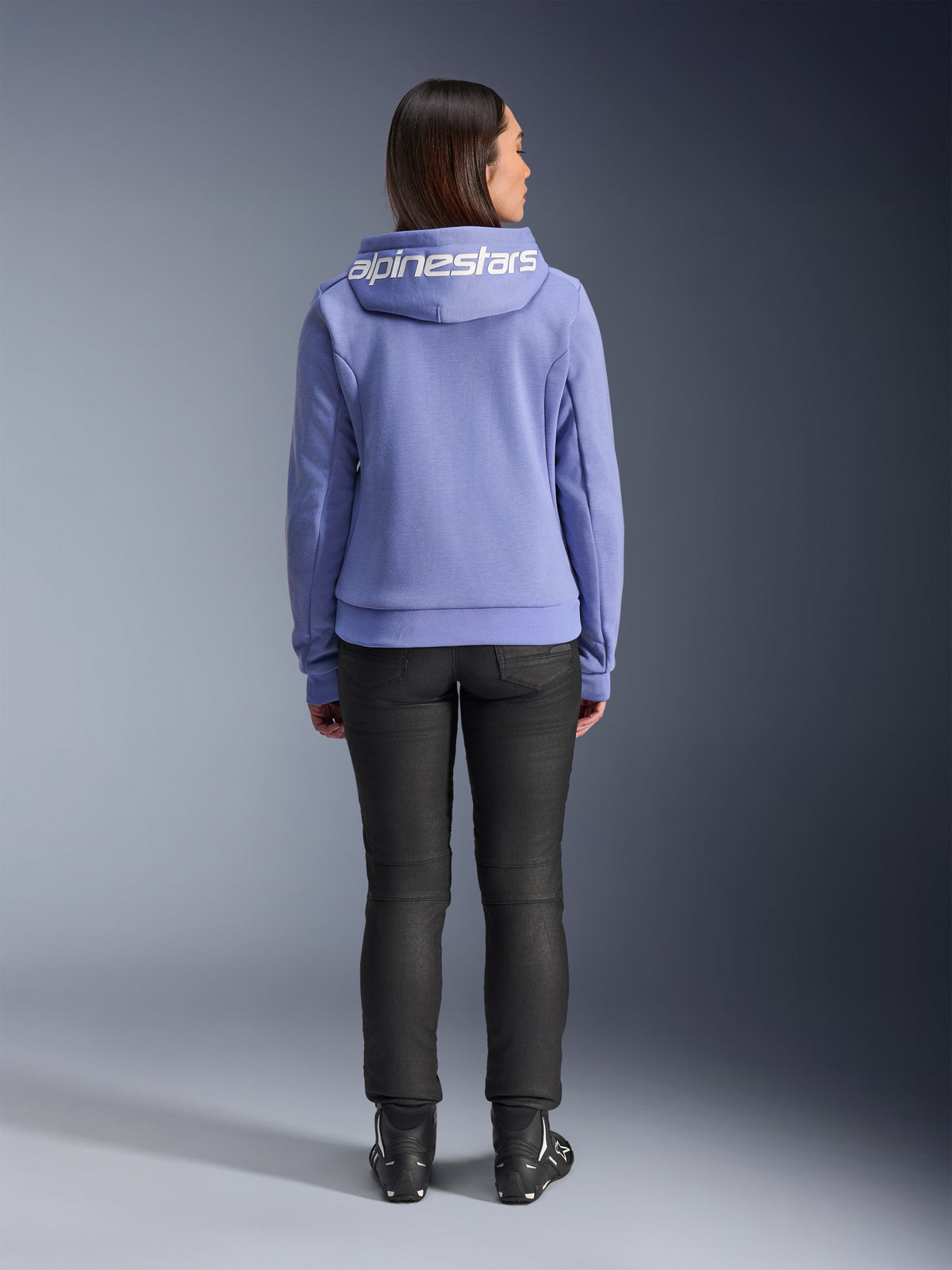 Alpinestars Stella Chrome V2 Sport Hoodie, azul violeta, vista posterior de una mujer con la sudadera que presenta el logotipo blanco de Alpinestars en la capucha, combinada con pantalones negros y botas de moto, ropa de moto urbana de estilo casual