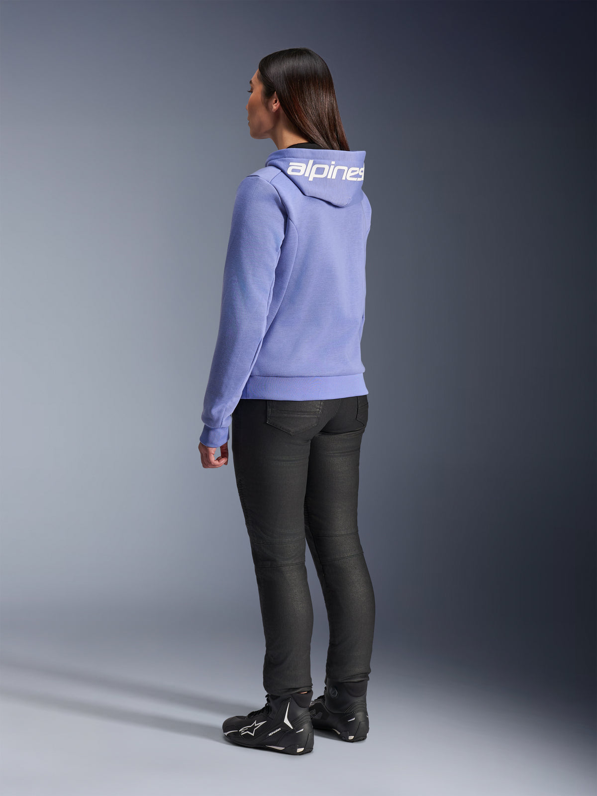 Sudadera deportiva Alpinestars Stella Chrome V2 para mujer, azul violeta, vista posterior que muestra el logotipo blanco en la capucha, usada con pantalones de montar negros y botas de moto negras, equipamiento casual de moto urbana