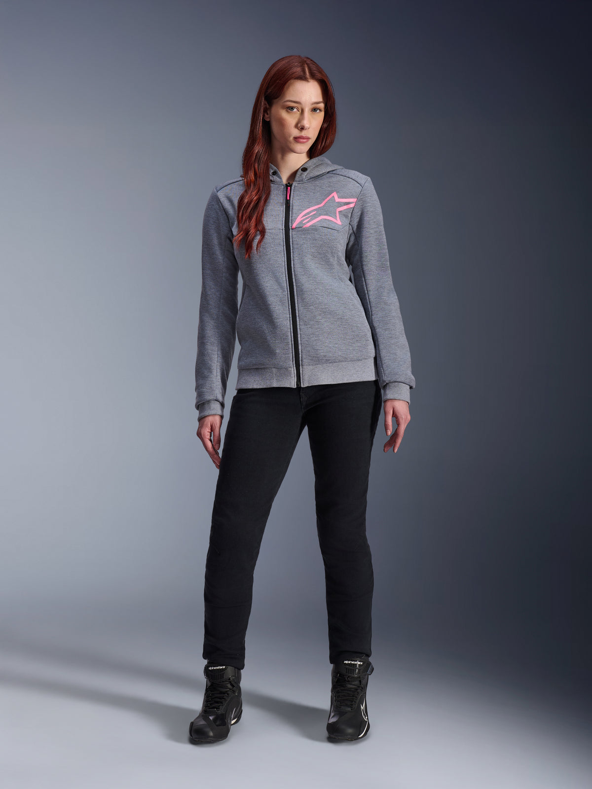 Alpinestars Stella Chrome V2 Sport Hoodie, gris jaspeado rosa flúor, vestida por una mujer con pantalones negros y botas, con el logotipo de la estrella Alpinestars en rosa en el pecho, equipamiento urbano para moto con cremallera completa, forro polar técnico ligero con un ajuste atlético casual.