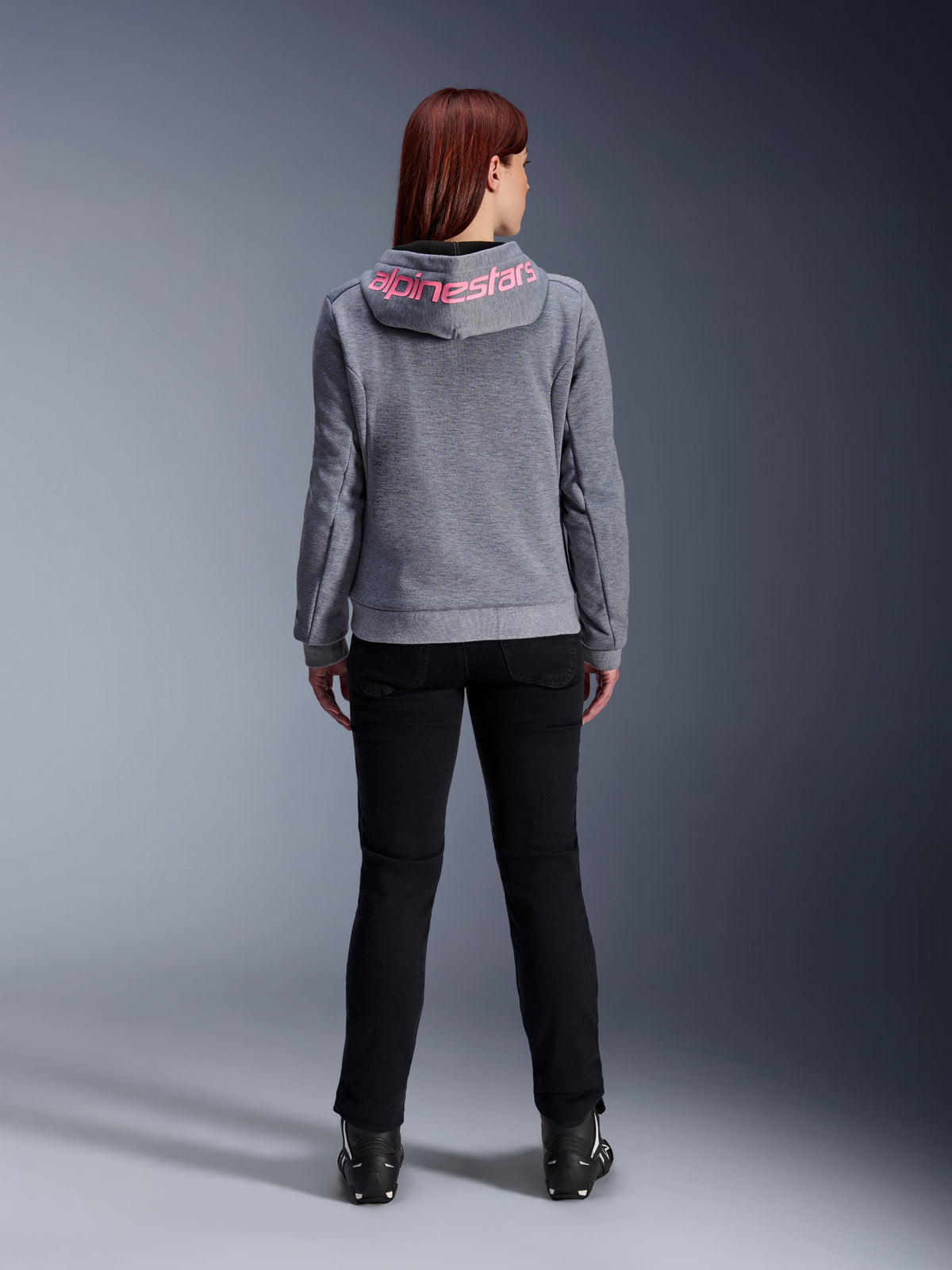 Alpinestars Stella Chrome V2 Sport Hoodie, sudadera de moto para mujer, gris jaspeado y rosa flúor, vista posterior que muestra el logotipo rosa de Alpinestars en la capucha, combinada con pantalones de conducción negros y botas, ropa de moto casual para mujer