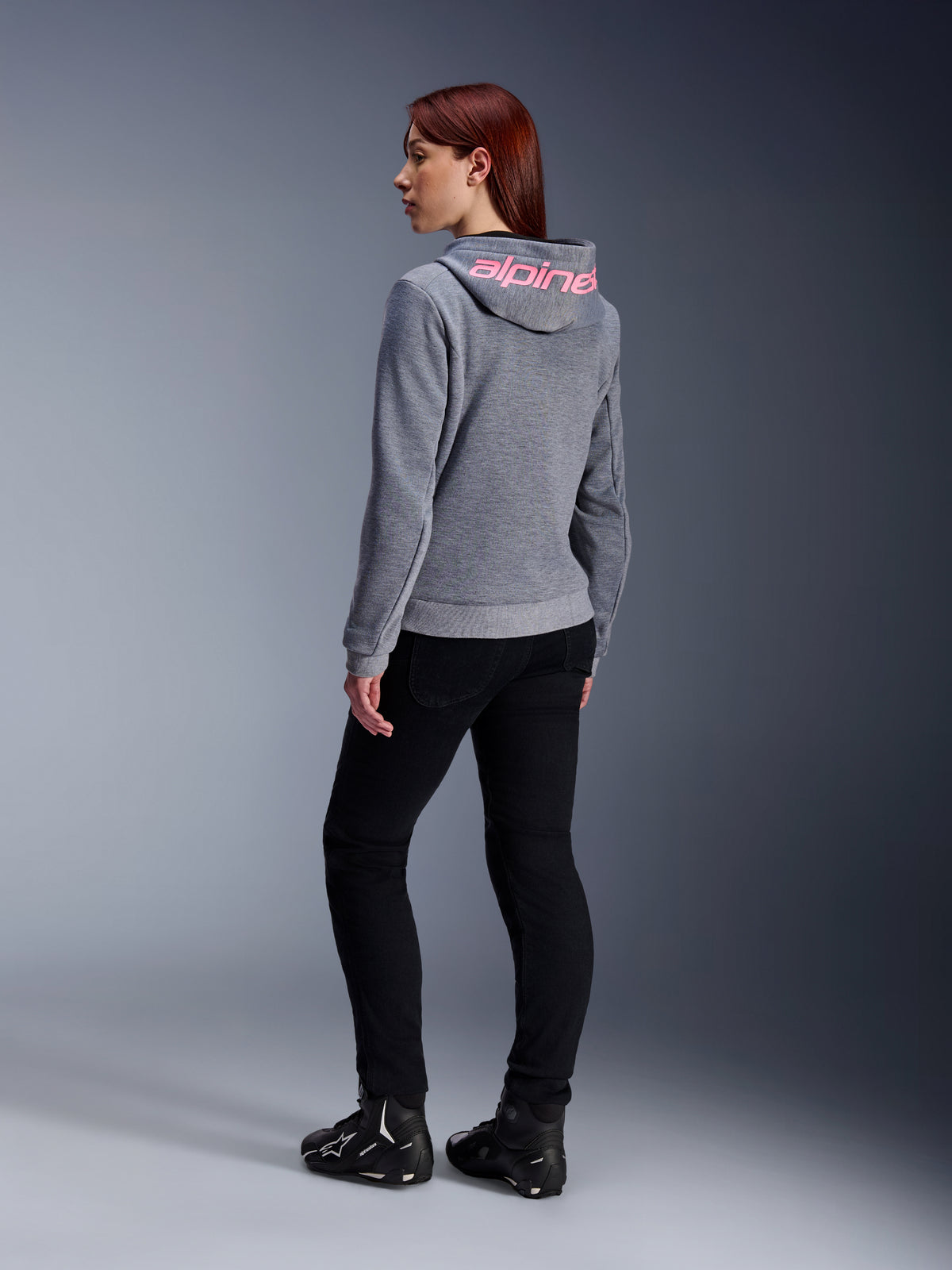 Alpinestars Stella Chrome V2 Sport Hoodie, sudadera de moto para mujer, gris jaspeado con logotipo rosa flúor en la capucha, vista trasera, combinada con pantalones negros y calzado de moto, estilo urbano casual con protección para motociclistas