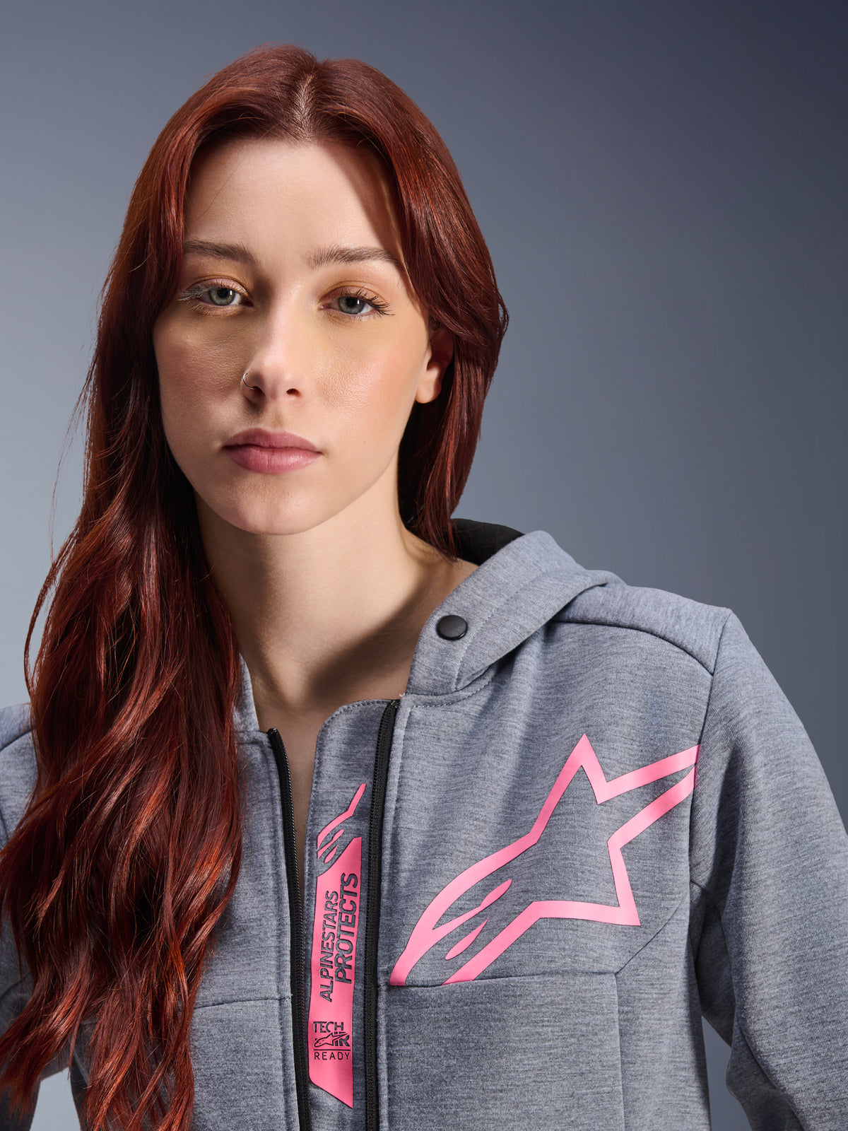Mujer con la sudadera Alpinestars Stella Chrome V2 Sport Hoodie, gris jaspeado y rosa flúor, primer plano que muestra el logotipo de la estrella rosa de Alpinestars y la marca Tech-Air Ready, ropa urbana para moto