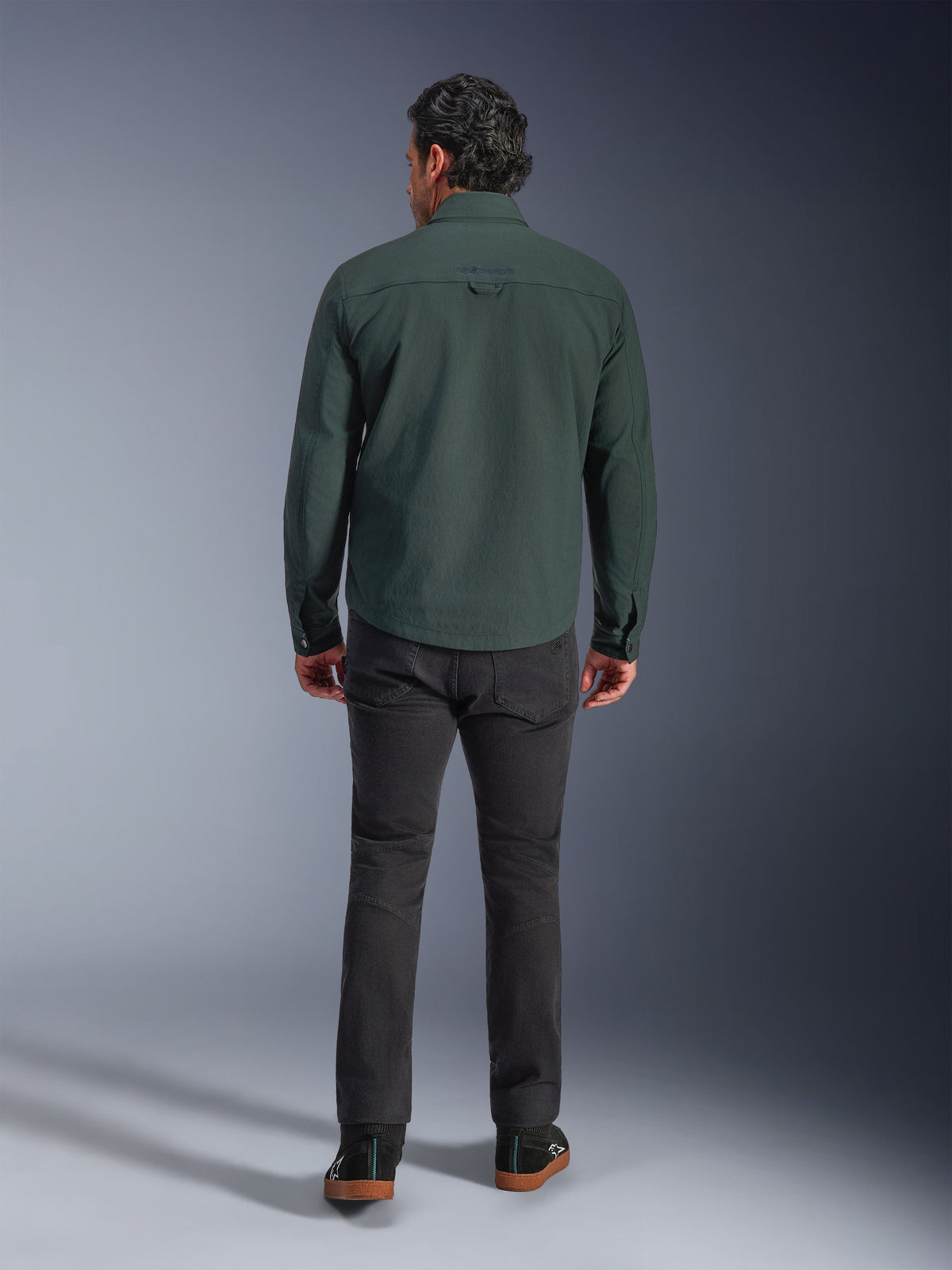Alpinestars Bryon Water Resistant Overshirt in Dark Emerald, back view on model, paired with dark grey motorcycle pants and black riding shoes, showcasing a classic workwear-inspired design with a slightly longer back for riding coverage.