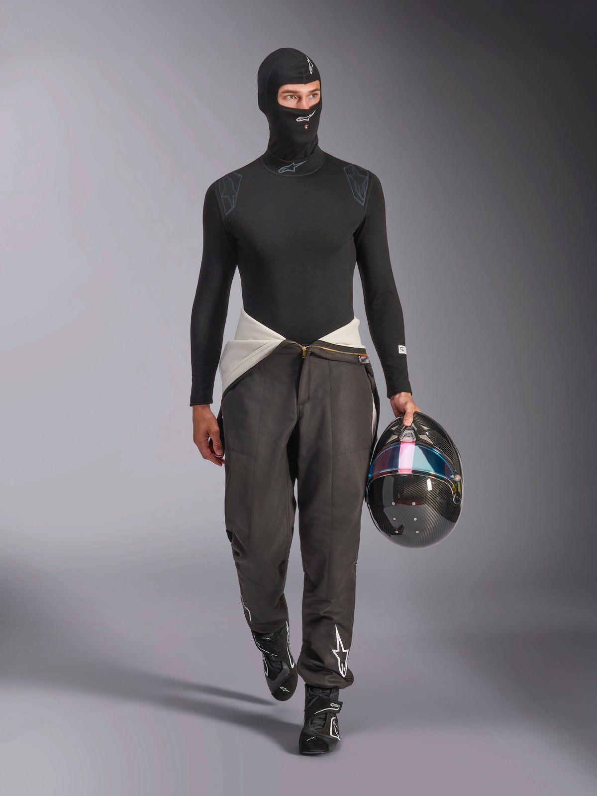 Alpinestars ZX Evo V3 Balaclava, negro, usado por un modelo con un conjunto completo de capas base para carreras, que incluye una parte superior de manga larga a juego y pantalones de carreras grises, sosteniendo un casco de fibra de carbono con visera iridiscente, equipamiento profesional para automovilismo