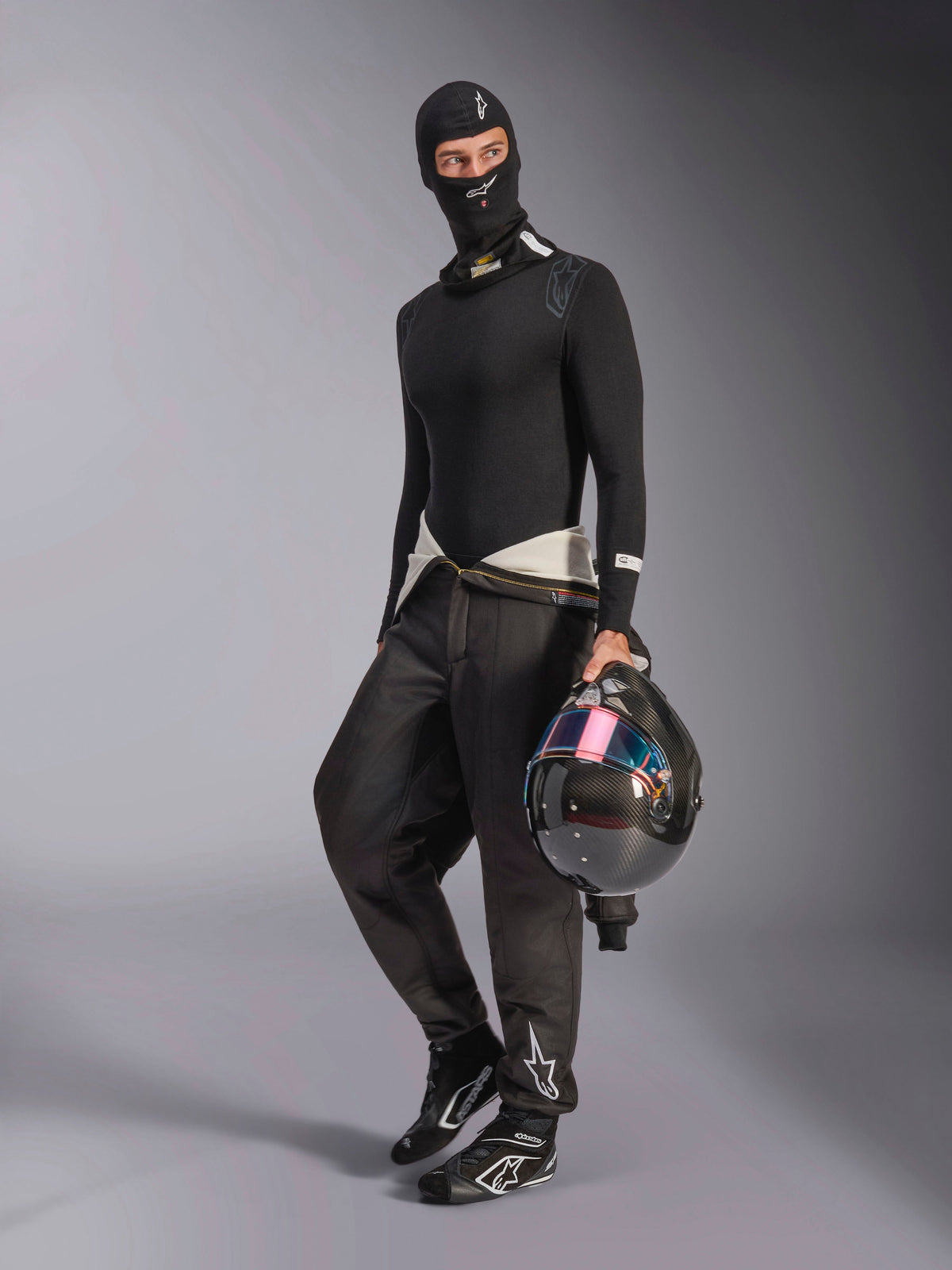 Pasamontañas Alpinestars ZX Evo V3, negro, usado por un piloto con equipo completo de capas base para automovilismo, con el logo blanco en la frente, sosteniendo un casco de fibra de carbono con visera iridiscente, ropa profesional de automovilismo resistente al fuego.