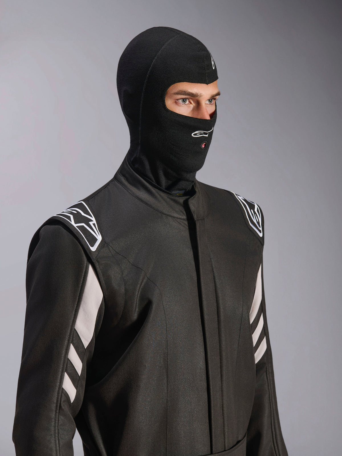 Pasamontañas Alpinestars ZX Evo V3, negro, sotocasco ignífugo para automovilismo, usado por un modelo con un traje de carreras negro con detalles blancos, incluye el logotipo de Alpinestars en la barbilla, puerto para tubo de hidratación y equipo de protección profesional para deportes de motor