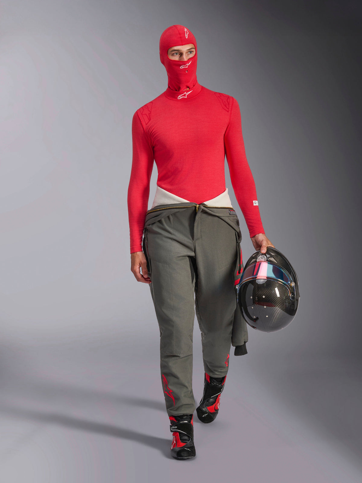 Piloto de carreras con pasamontañas Alpinestars ZX Evo V3 en rojo, capa base de manga larga roja a juego, pantalones de carreras grises, botas de carreras negras y rojas, sujetando un casco de fibra de carbono, conjunto de equipo profesional para automovilismo