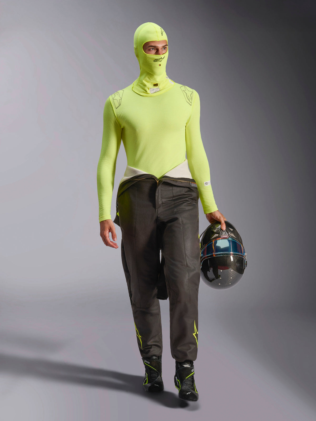 Piloto de carreras con pasamontañas Alpinestars ZX Evo V3 en Amarillo Fluo, camiseta interior de manga larga a juego, pantalones de carreras oscuros y botas de carreras negras, sujetando un casco de fibra de carbono, equipo profesional de automovilismo