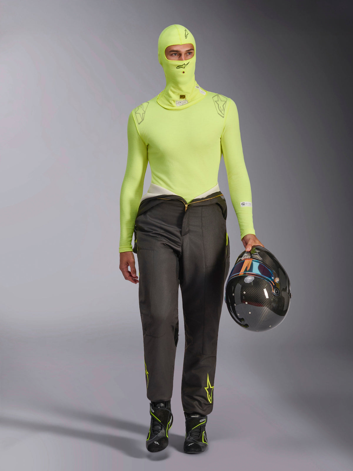 Pasamontañas Alpinestars ZX Evo V3 en Amarillo Fluo, usado con camiseta de capa base de manga larga a juego y pantalones de carreras gris oscuro, modelo masculino sosteniendo un casco de fibra de carbono, equipo profesional de automovilismo