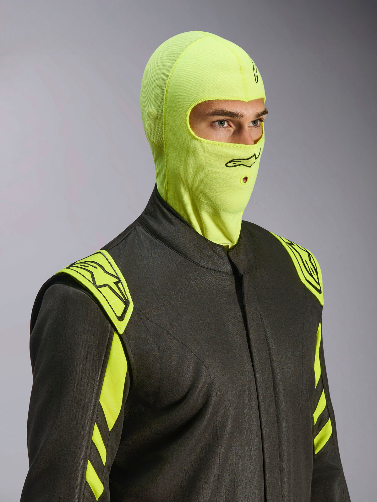 Modelo masculino con pasamontañas Alpinestars ZX Evo V3 en Yellow Fluo, sotocasco de competición resistente al fuego, combinado con un traje de automovilismo gris oscuro con detalles en amarillo neón, con logotipo de Alpinestars negro y apertura para tubo de hidratación, equipo de protección profesional para automovilismo