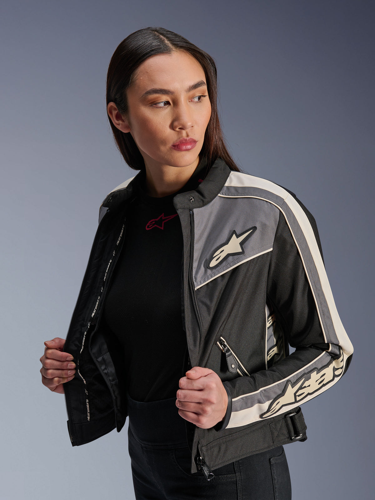 Alpinestars Stella T-Dyno WR para mujer, chaqueta textil de moto, negro, gris oscuro y crudo, diseño de inspiración vintage con franjas deportivas y logotipos en el pecho y las mangas, lucida por una modelo, equipamiento para conducción urbana