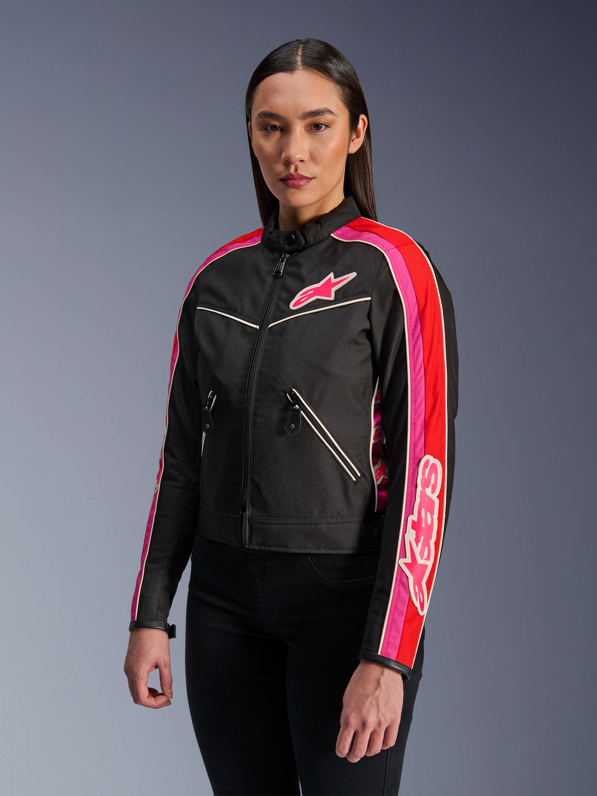 Mujer vistiendo la chaqueta Alpinestars Stella T-Dyno WR en color negro, rojo brillante y fucsia, chaqueta de moto de tejido textil con un ajuste de motociclista de inspiración vintage, con vibrantes franjas fucsias y rojas en las mangas y un logotipo en el pecho, combinada con pantalones negros.