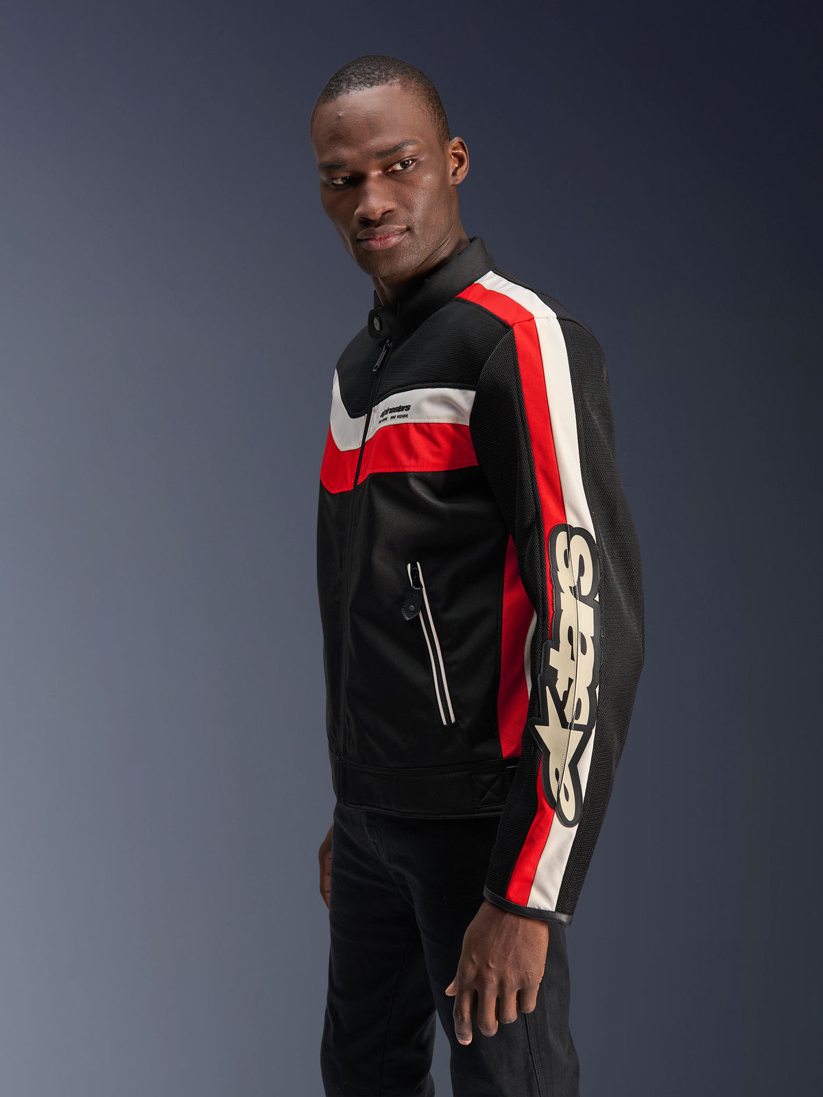 Modelo masculino con la chaqueta Alpinestars T-Dyno Air en negro, rojo brillante y crudo, chaqueta de moto textil de inspiración vintage con paneles de malla 3D, rayas llamativas en rojo y blanco hueso, y logotipo de Alpinestars destacado en la manga