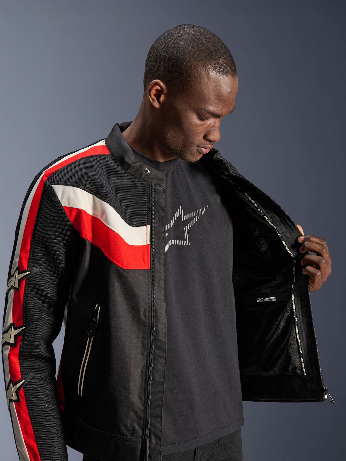 Chaqueta Alpinestars T-Dyno Air, negro, rojo brillante y ecru, modelo masculino abriendo la chaqueta para mostrar el forro interno y el compartimento impermeable, chaqueta textil de inspiración vintage con paneles de malla 3D y logos de estrellas, equipamiento moderno para motociclismo