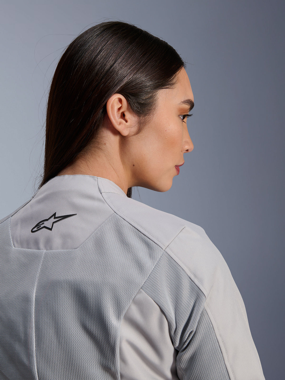 Mujer vistiendo la chaqueta Stella C-1 Air de Alpinestars en color plateado, vista lateral-posterior de primer plano que muestra el logotipo negro de Alpinestars, paneles de ventilación de malla técnica y un diseño de moto urbano, limpio y contemporáneo.