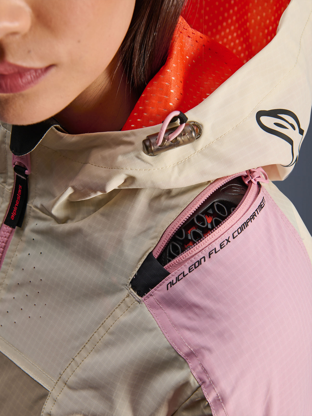 Primer plano de la chaqueta Alpinestars Molly Air en Sand Pink Almond, destacando el compartimento con cremallera en el hombro para protectores Nucleon Flex, el forro de malla naranja brillante de la capucha y la construcción duradera en tejido ripstop.