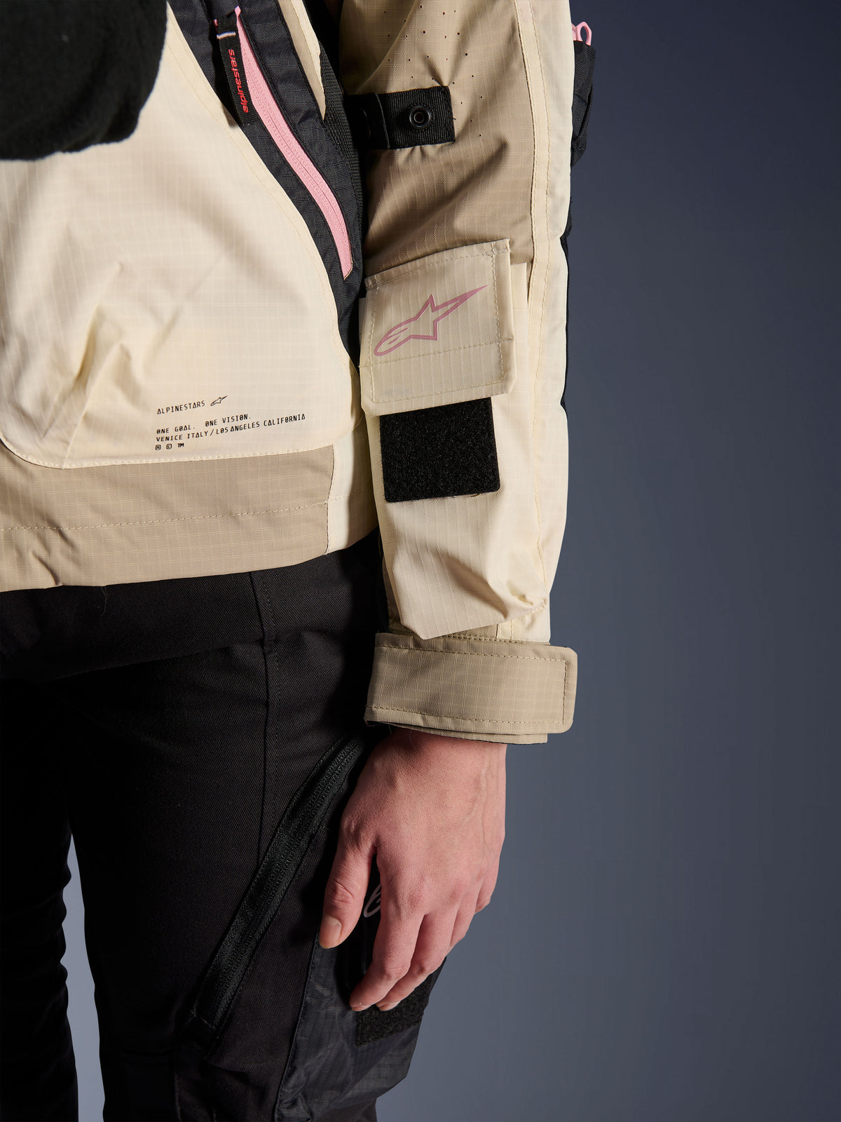 Chaqueta Alpinestars Molly Air, chaqueta de moto urbana para mujer, Sand Pink Almond, vista lateral en primer plano que muestra el bolsillo de la manga con logotipo rosa, parche de velcro negro y texto de la misión de la marca en la parte inferior de la espalda, material ripstop ligero