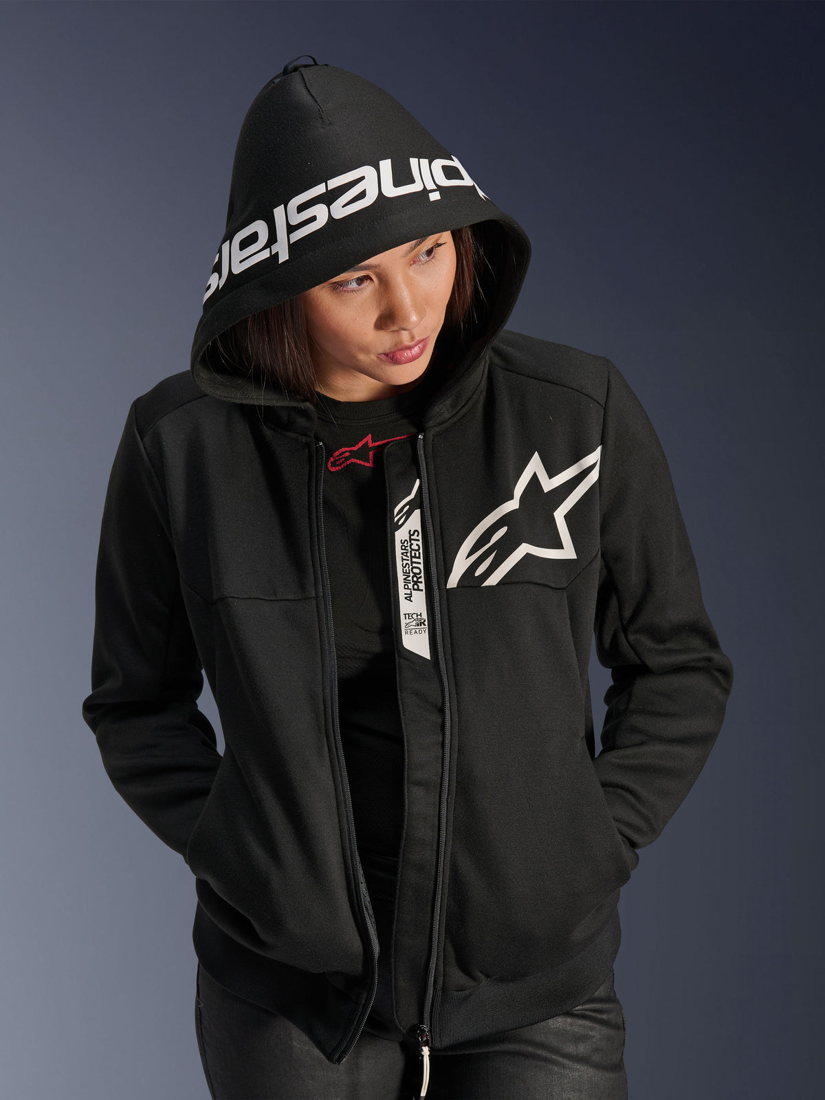 Sudadera deportiva Alpinestars Stella Chrome V2 para mujer, negro y blanco, sudadera para moto con un logotipo de estrella blanca en el pecho y marca Alpinestars en el borde de la capucha, mujer con la capucha puesta, estilo deportivo urbano casual con detalles Tech-Air Ready.