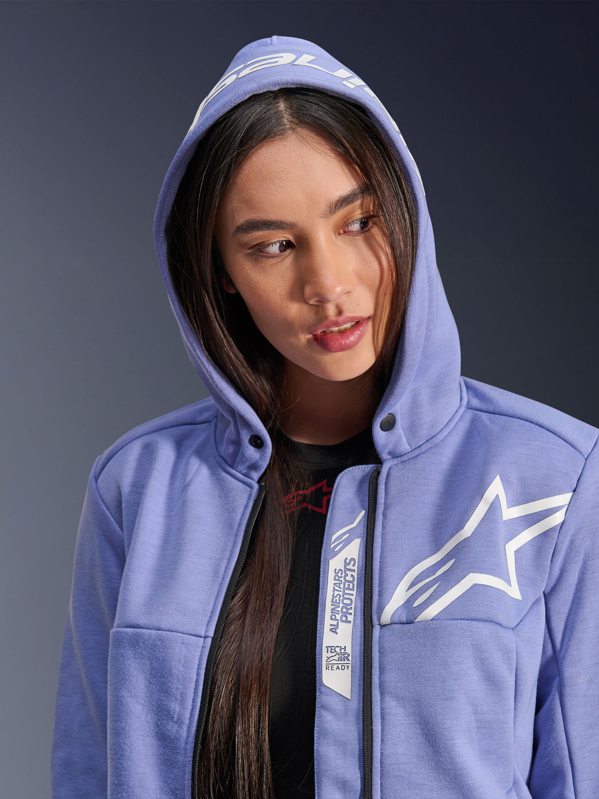 Sudadera deportiva Alpinestars Stella Chrome V2, sudadera de moto para mujer, color violeta azulado con logo de estrella blanco, con marcas 'Alpinestars Protects' y 'Tech-Air Ready' junto a la cremallera, diseño urbano casual con la capucha puesta.