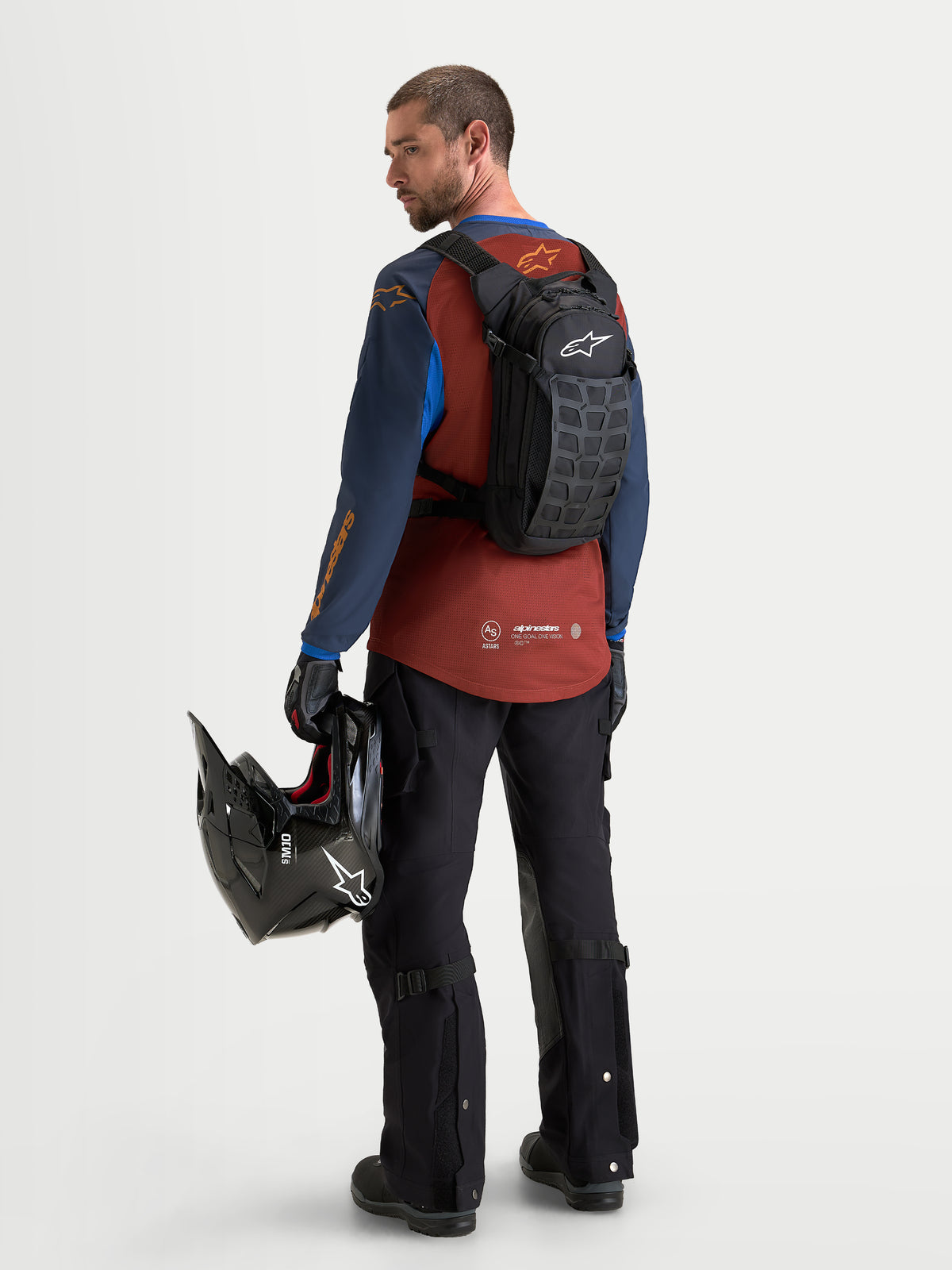Alpinestars Mochila AT-9, mochila de motocross en negro y gris oscuro, vista trasera de un modelo con equipamiento de equitación y sosteniendo un casco