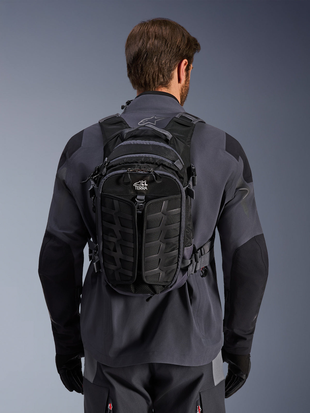 Mochila Alpinestars AT-15, mochila de moto para aventura, Negro Deep Gray, llevada por una persona sobre una chaqueta técnica de moto gris, vista trasera con el logotipo 'ALL TERRA', diseño ergonómico con paneles texturizados reforzados y correas seguras.