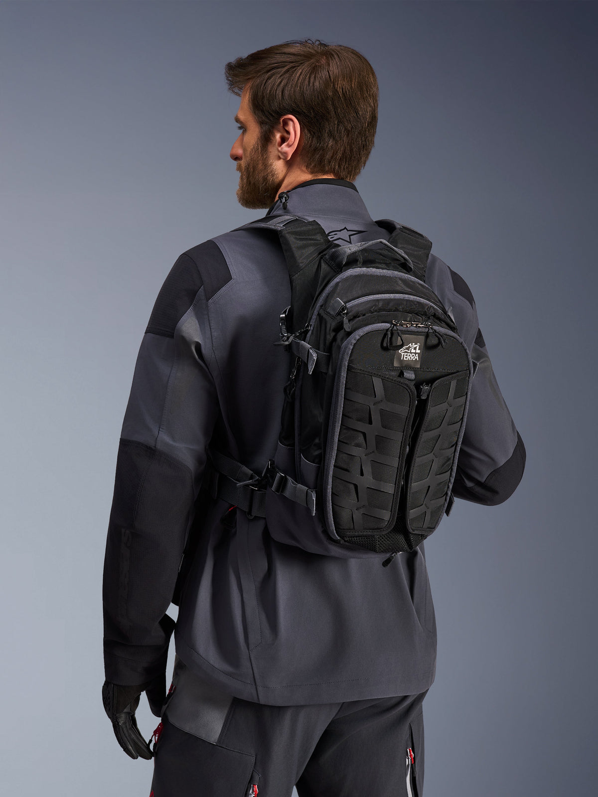 Mochila Alpinestars AT-15, mochila de aventura para motocicleta, negra y gris oscuro, llevada por un piloto con una chaqueta gris, con un diseño técnico con cinchas MOLLE y marca 'TERRA', equipamiento ergonómico para off-road