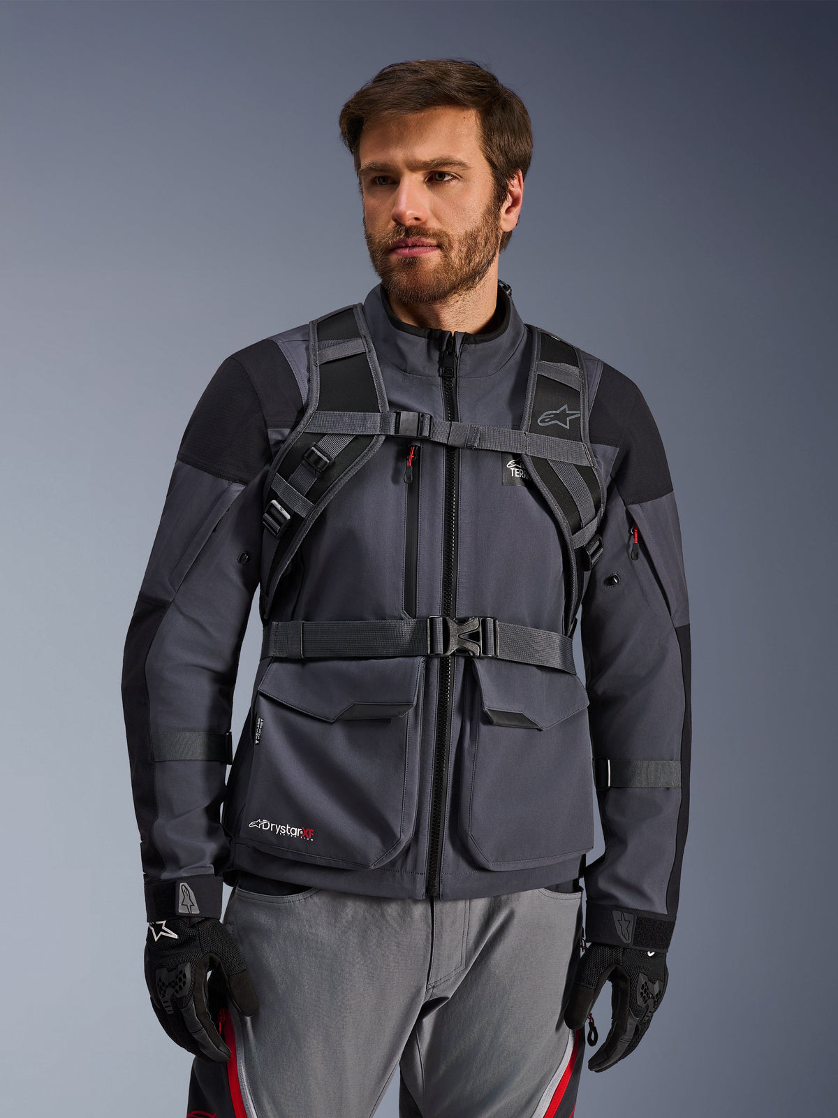 Sistema de arnés de la mochila Alpinestars AT-15, Black Deep Gray, vista frontal usada por un motorista sobre una chaqueta de moto gris, con cierre magnético en el pecho y correa de cintura segura, combinada con guantes negros para motociclismo de aventura