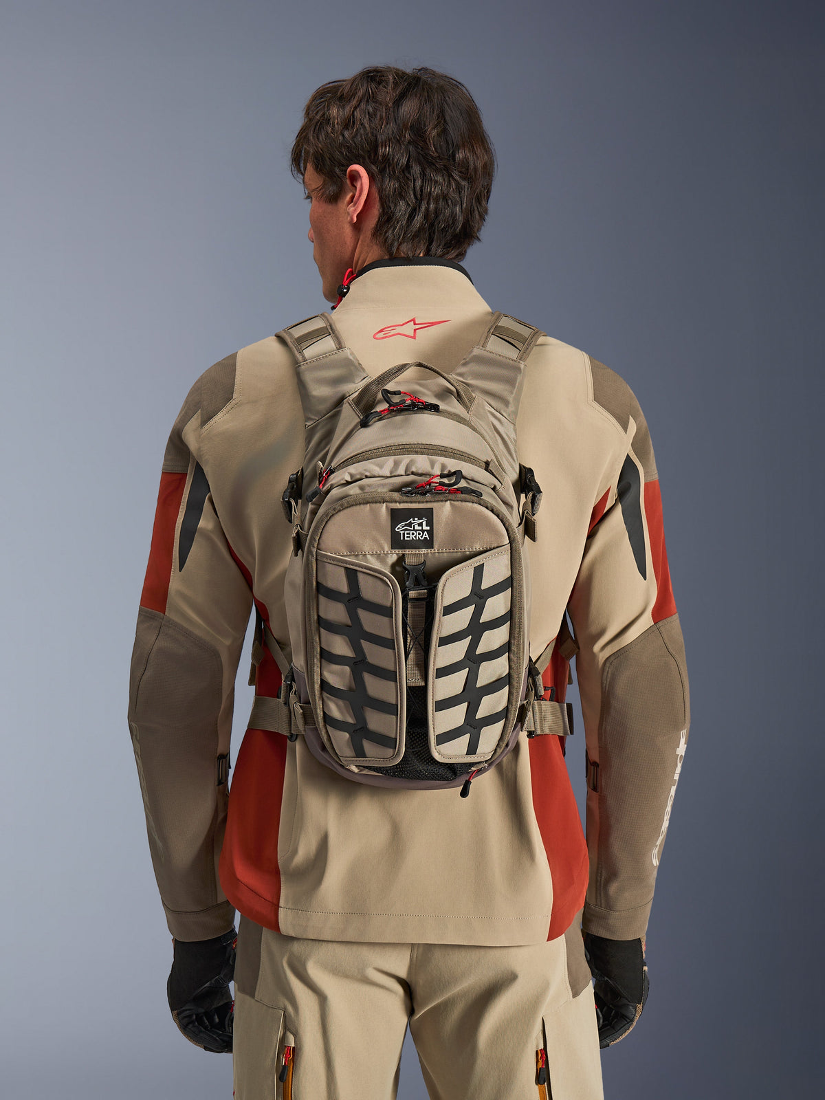 Alpinestars AT-15 Back Pack, Aluminum Walnut, mochila técnica de aventura para motociclismo usada por un piloto con chaqueta beige y roja, con patrón de dibujo de neumático, logotipo TERRA, tiradores de cremallera rojos y correas ajustables