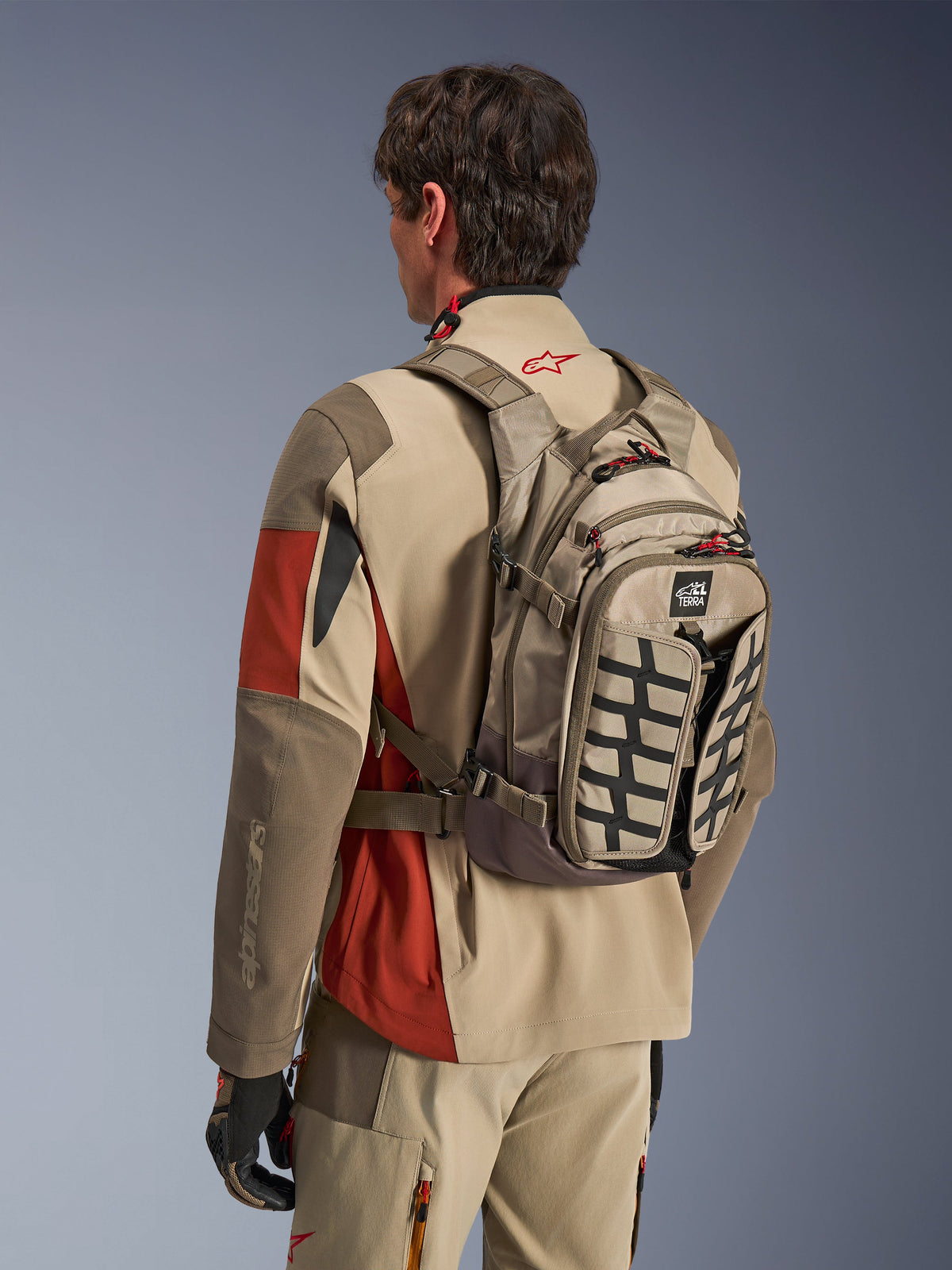 Mochila Alpinestars AT-15 en color Aluminum Walnut, mochila técnica de motociclismo portada por un piloto con equipamiento de aventura a juego, con confección textil en tonos arena y marrón, detalles estructurales en negro y tiradores de cremallera rojos, diseñada para conducción off-road y de aventura.