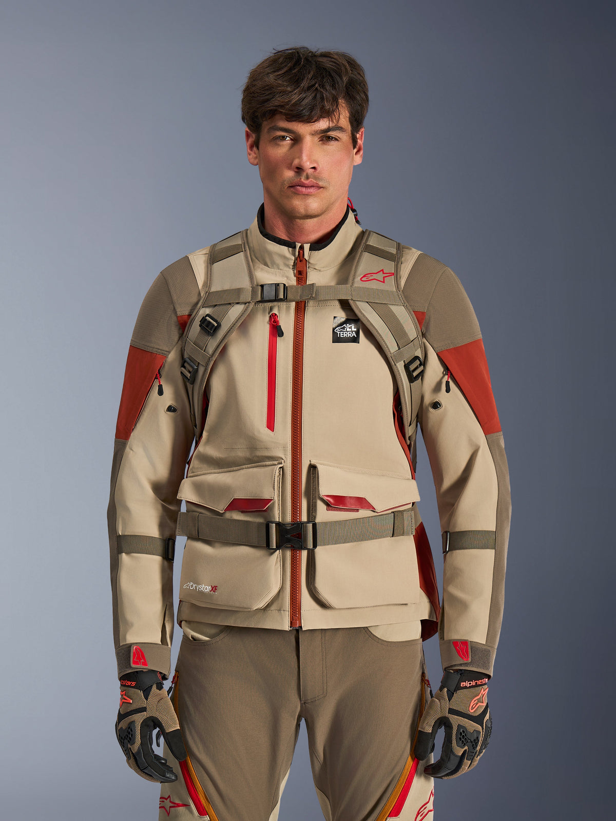 Piloto de aventura vistiendo la mochila Alpinestars AT-15 en Aluminum Walnut, vista frontal que muestra las correas del pecho y de la cintura con cierre magnético, combinada con chaqueta y pantalones de moto de aventura en color tostado y rojo, conjunto de equipamiento técnico para off-road
