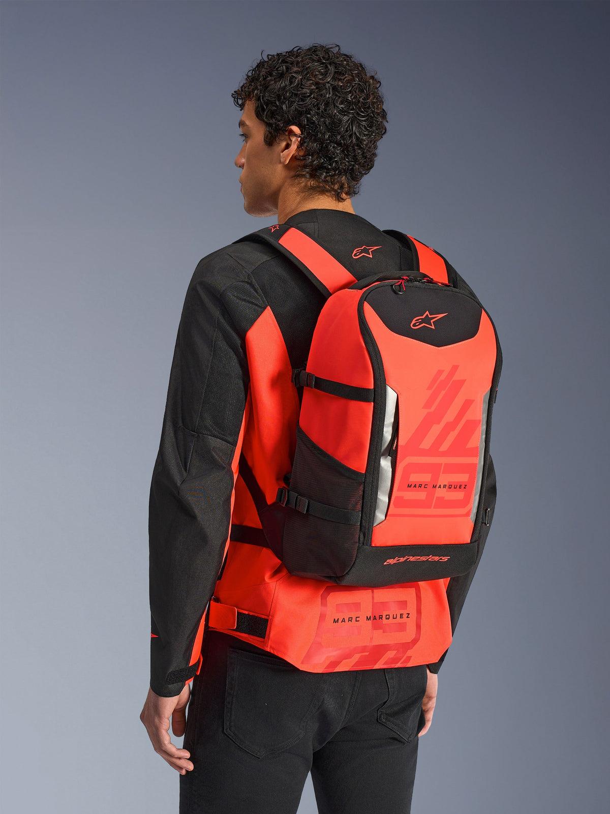 Alpinestars MM93 AMP3 Backpack, Technical Motorcycle Backpack, Red Fluo and Black, worn by a model from behind over a matching red and black jacket, featuring Marc Marquez '93' graphics, adjustable straps, and sleek sport-inspired design.