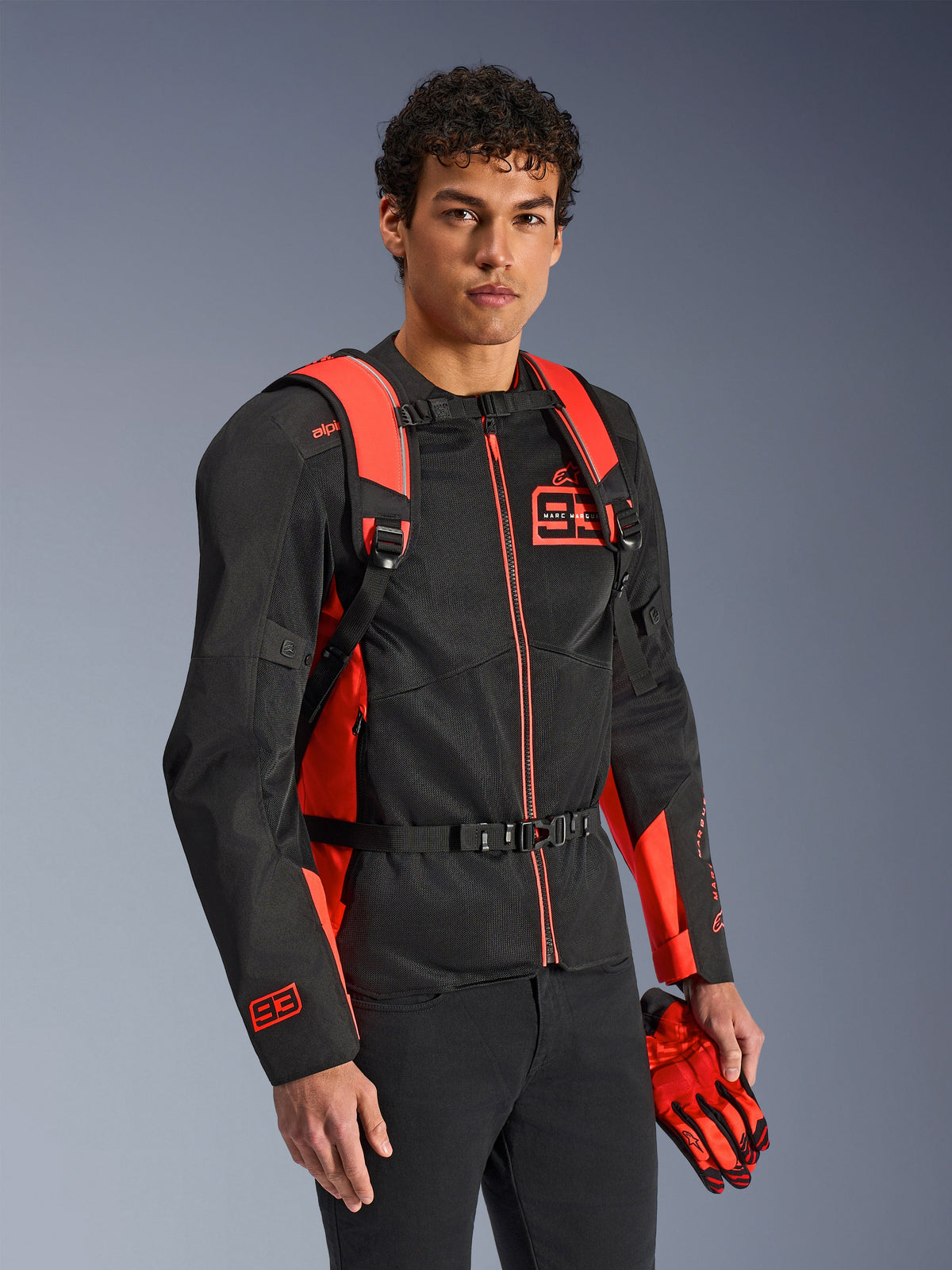 Alpinestars MM93 AMP3 Backpack, Red Fluo and Black, worn by a model with a matching black and red MM93 motorcycle jacket and gloves, front view highlighting the adjustable shoulder straps and chest buckle, technical racing-inspired gear.