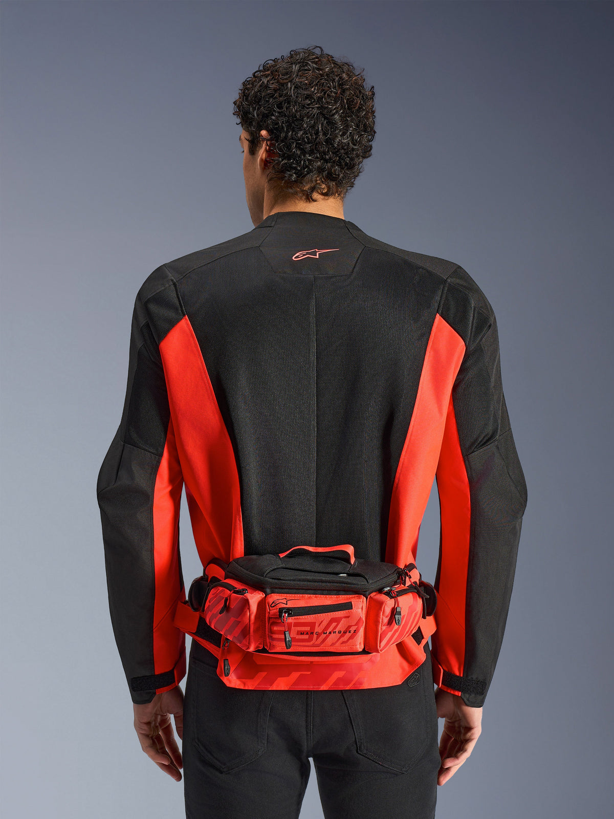 Riñonera Alpinestars MM93, roja y negra, usada sobre una chaqueta de moto negra y roja, con logotipos de Marc Márquez 93, cinturón ajustable y compartimentos con cremallera, accesorio funcional inspirado en la competición