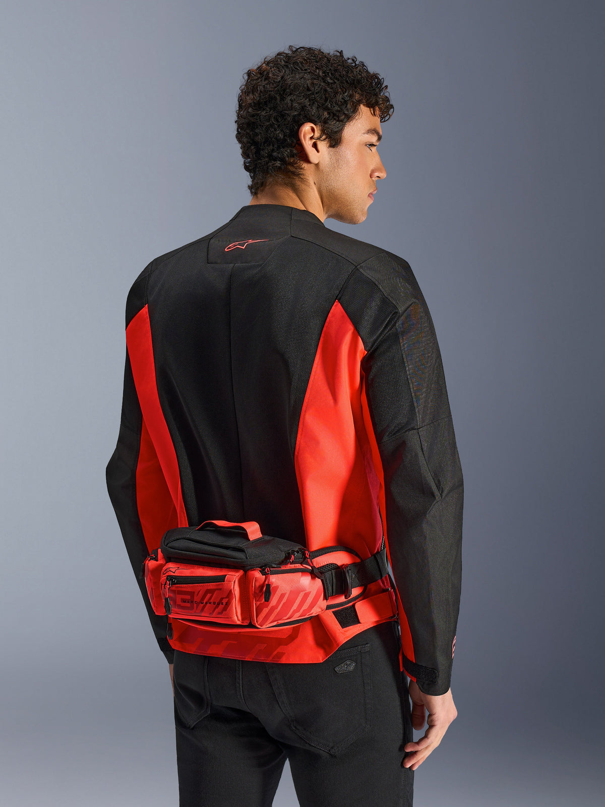 Alpinestars MM93 Waist Bag, riñonera de moto, rojo brillante y negro, usada sobre una chaqueta de motociclismo a juego en negro y rojo, con compartimentos con cremallera y marca Marc Marquez MM93, construcción textil duradera para motociclismo