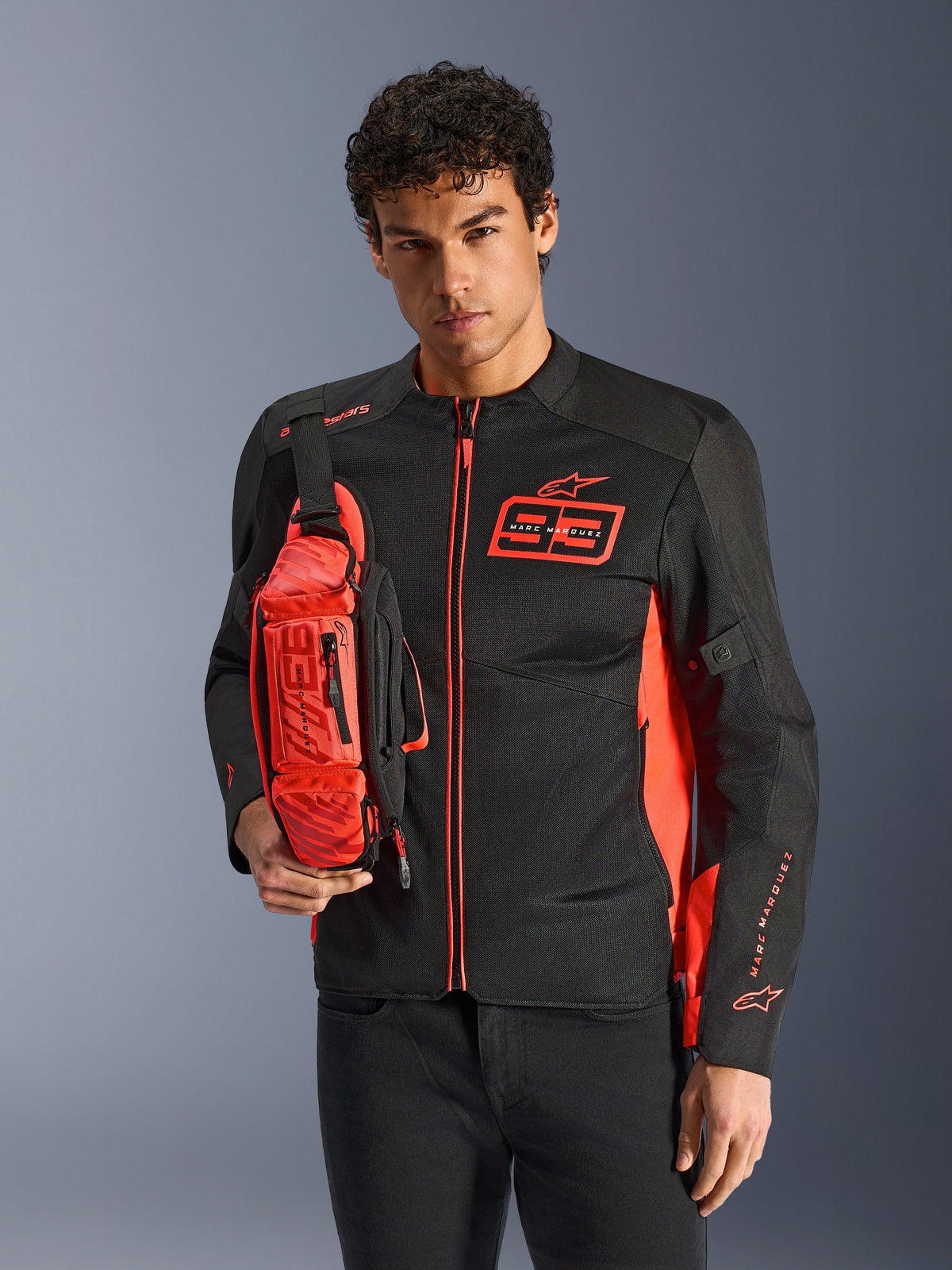 Hombre con la riñonera Alpinestars MM93, rojo flúor y negro, llevada como bandolera sobre una chaqueta de moto a juego en negro y rojo, con el logotipo de Marc Márquez 93 y diseño deportivo racing