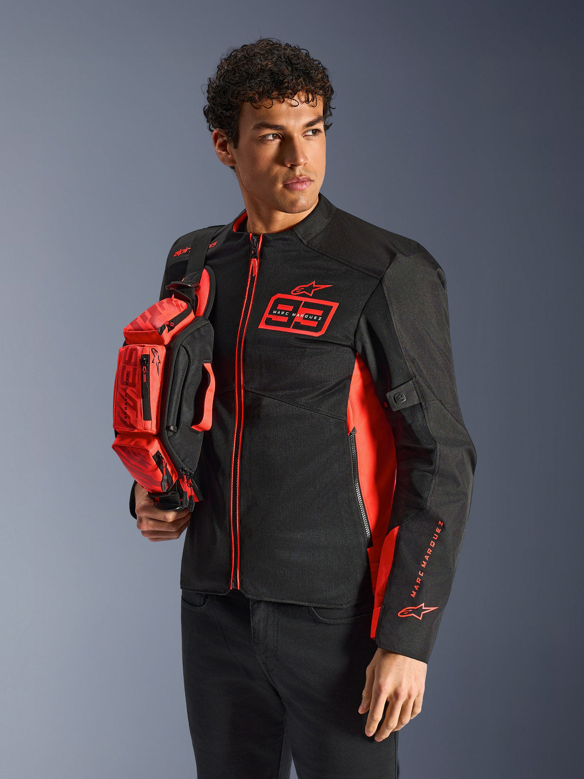 Alpinestars MM93 Waist Bag, rojo flúor y negro, sostenida por un modelo que viste una chaqueta de motociclista negra y roja a juego, con múltiples compartimentos con cremallera y el logotipo de Marc Márquez, equipamiento práctico para conducción deportiva