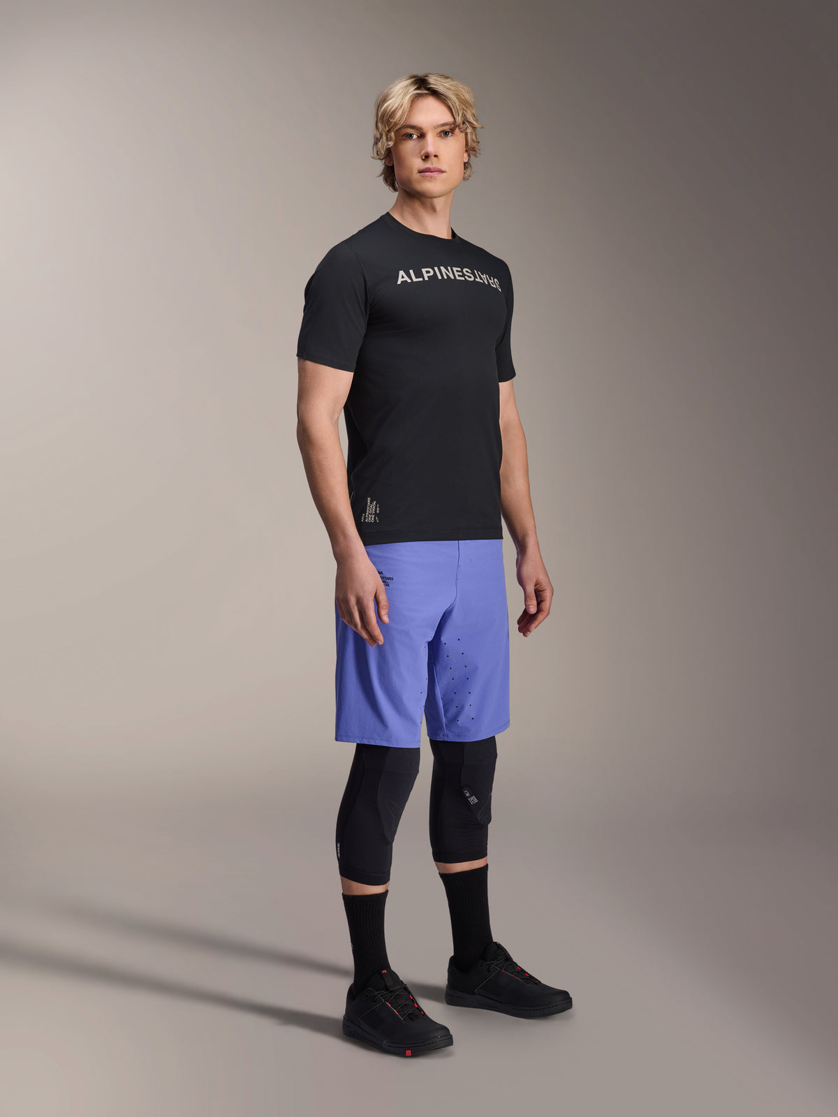 Male model wearing Alpinestars mountain bike apparel, featuring the black A-Aria Switch SS Jersey and Baja Blue A-Aria Lite Shorts, paired with black knee guards, black socks, and black mountain bike shoes in a full-body studio shot.
