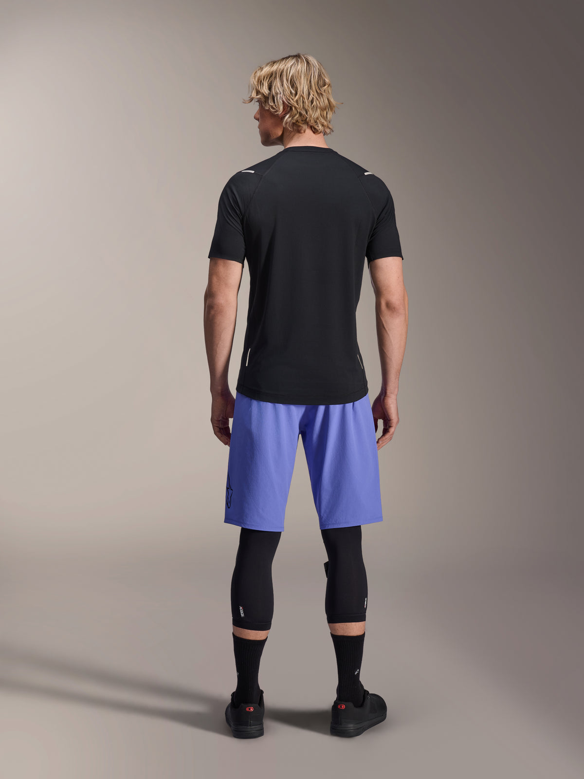 Jersey de manga corta Alpinestars A-Aria Switch, jersey de MTB negro, visto desde atrás, combinado con pantalones cortos de ciclismo de montaña azules y protección para las piernas negra, equipamiento de ciclismo de alto rendimiento para trail